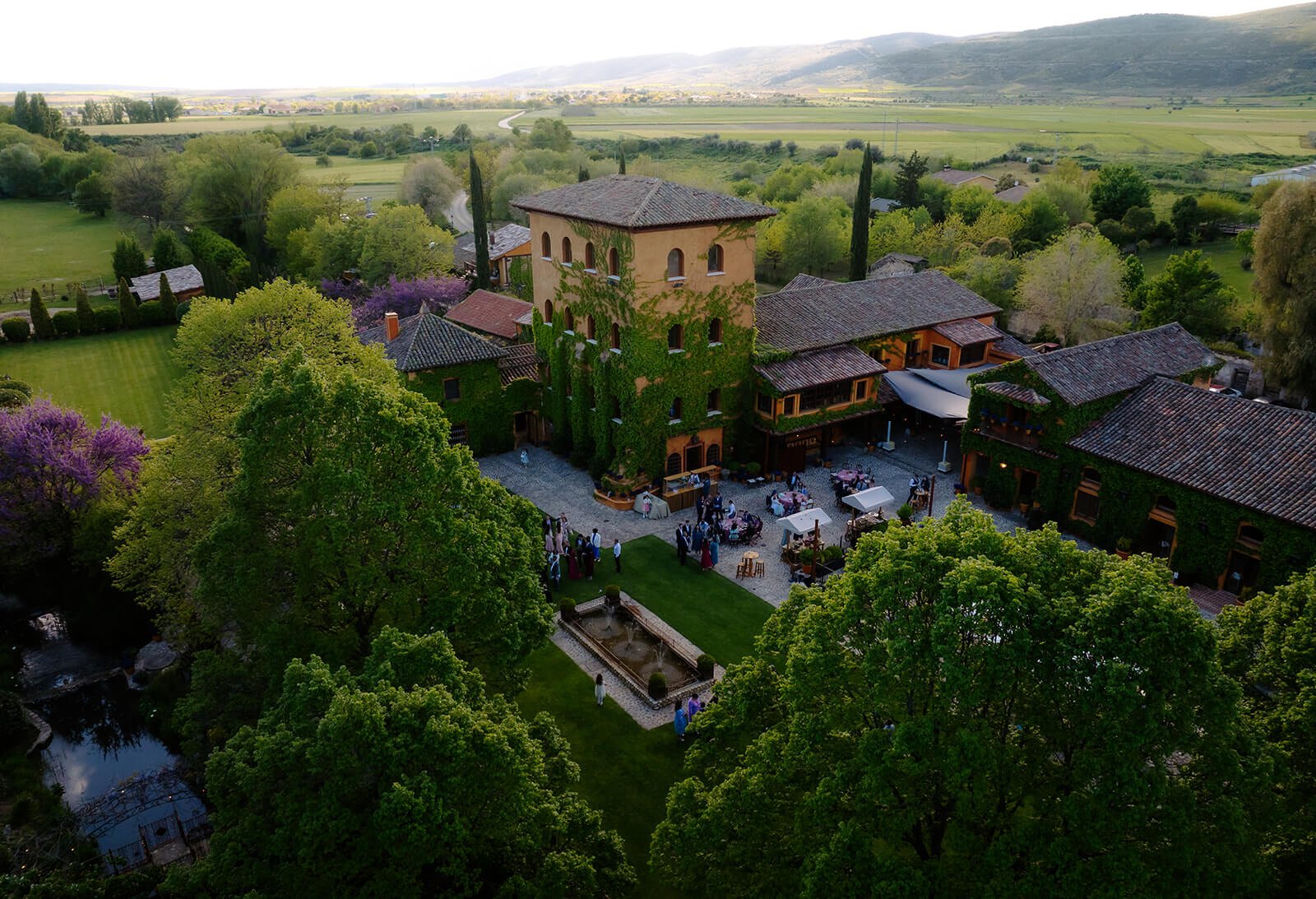 Toma Aérea de Finca para Bodas en la Sierra de Madrid con Jardines y Montañas Vista aérea de la finca de bodas en Madrid con el edificio principal, extensos jardines y los invitados disfrutando del cóctel de bienvenida.