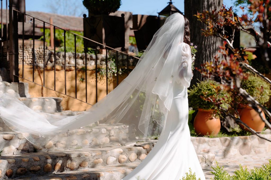 Retrato de la novia de espaldas subiendo la escalera de piedra en los jardines, con su velo largo ondeando al viento. Boda en Madrid.