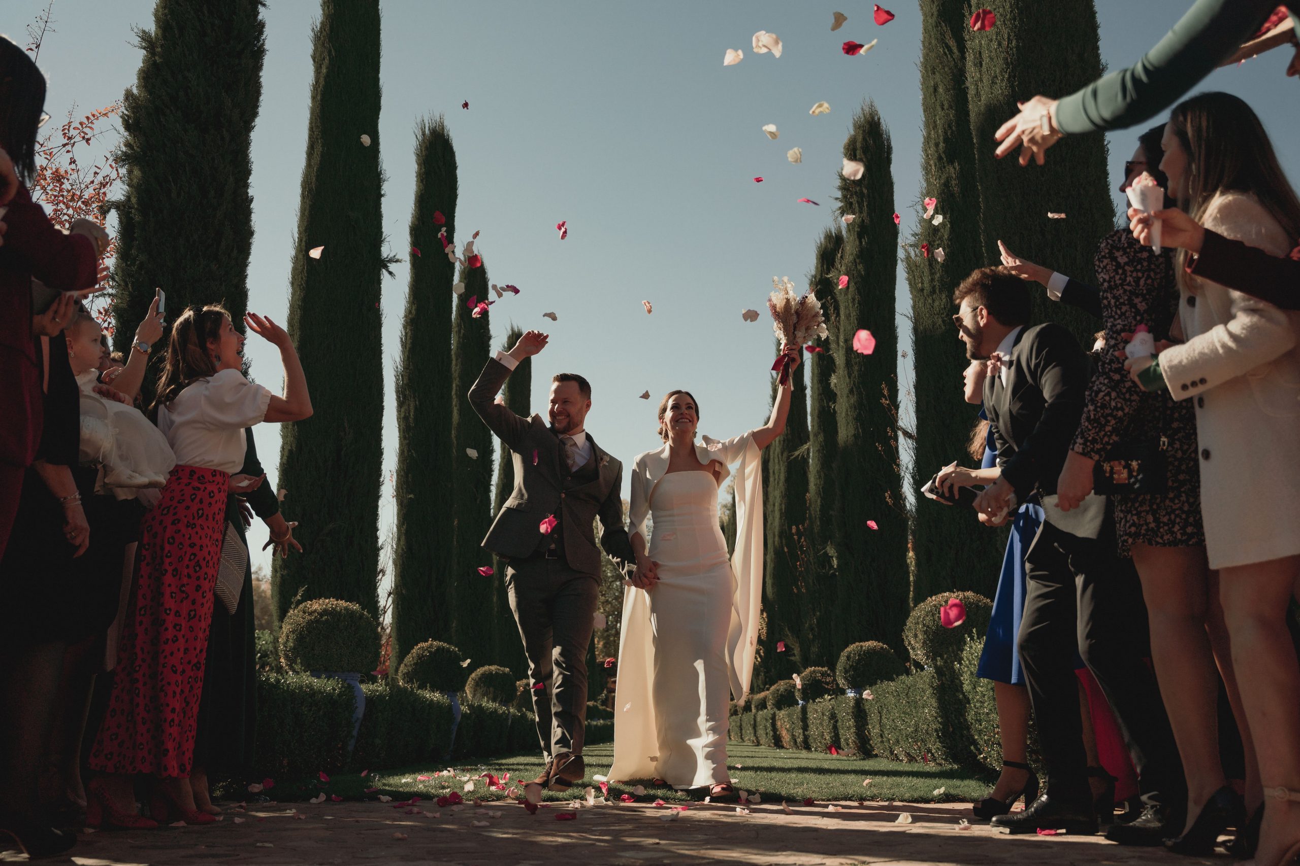 Momentos Inolvidables: Lanzamiento de Pétalos a los Novios en Boda de Otoño Novios saliendo de la ceremonia con los brazos en alto, mientras invitados lanzan arroz y pétalos de flores. Fondo de cipreses en una boda de otoño en Madrid.