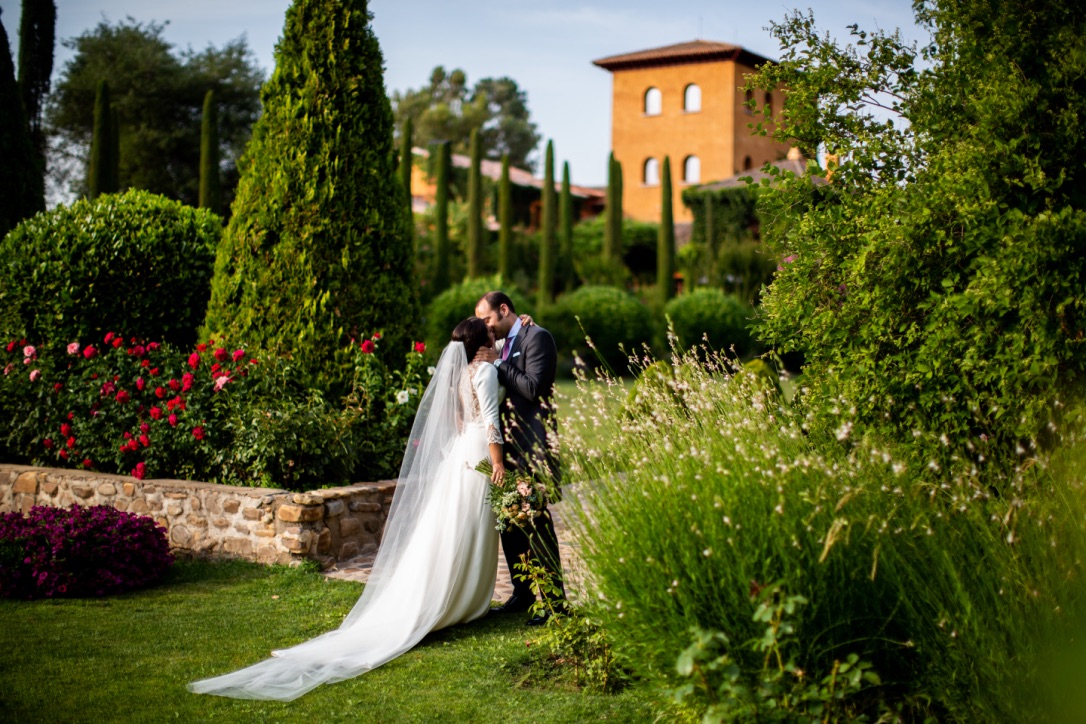 Pareja de boda en un jardín rodeado de vegetación y flores, con un edificio de tonos tierra al fondo.